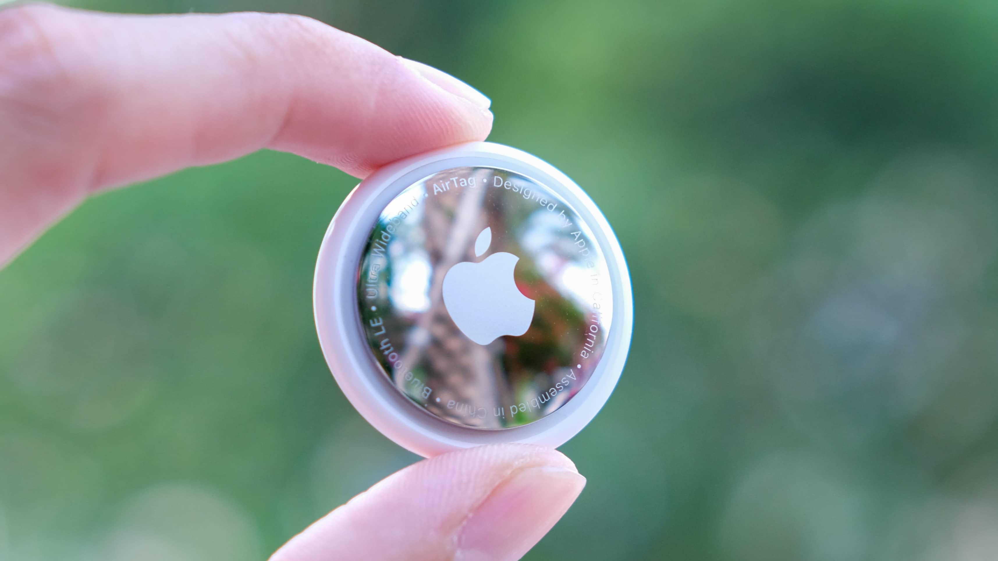 A closeup photograph of an Apple AirTag being held between a person's thumb and index finger, with the tracker's stainless steel back exposed to the camera