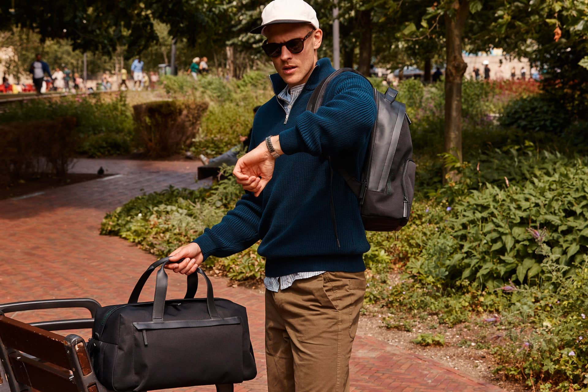 Young man standing in a park, telling the time on his watch while holding the handle of his Troubadour duffle bag sitting on a bench