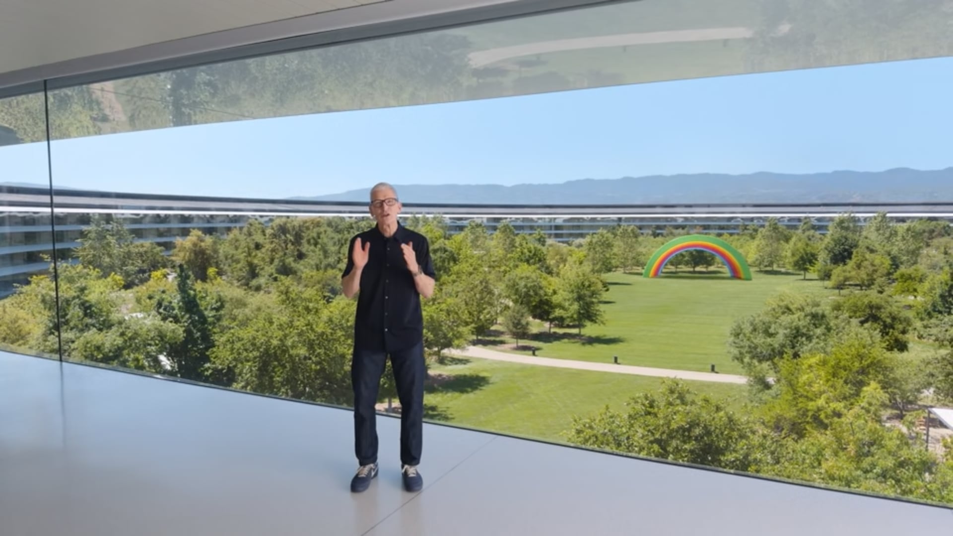 Tim Cook standing inside the Apple Park headquarters, with an outdoor rainbow-colored stage seen behind him through the glass walls.